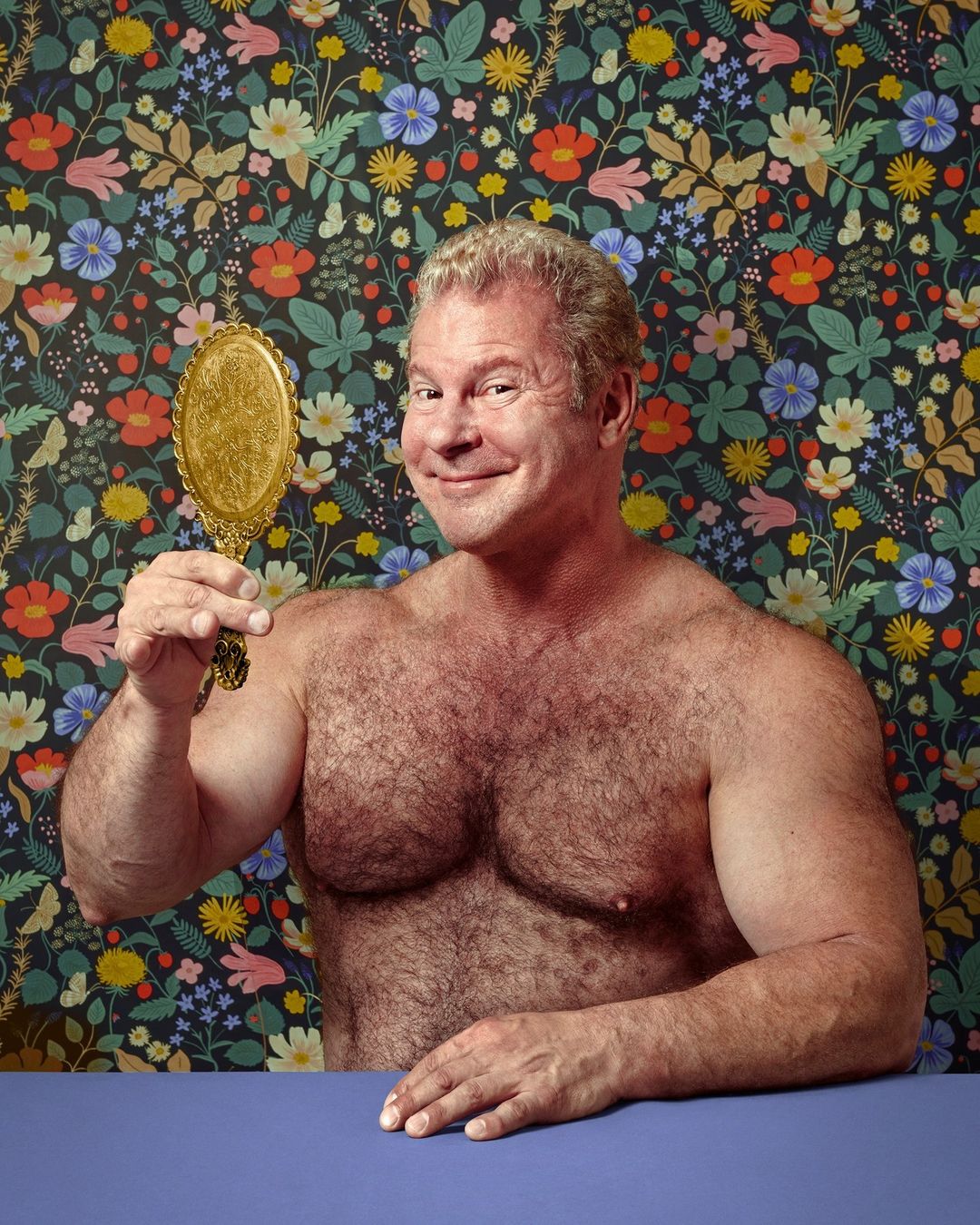 Adult man smiles while looking in a vintage gold hand mirror with a floral patterned background and blue table.